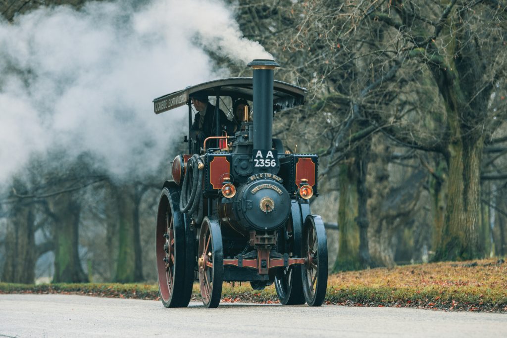 Vintage steam-powered vehicle in a rural autumn setting, surrounded by old trees.